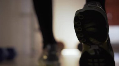 At the gym, a man in sneakers takes a few steps forward to the dumbbells, slows down, thinking next actions. He is in a fitness center where he is doing a workout. There are several dumbbells and a