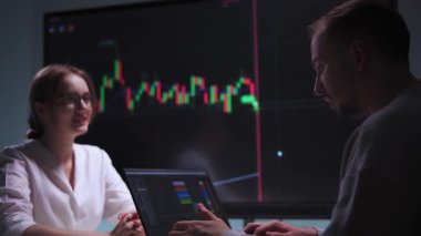 A man is sitting at a desk in an office, typing on a laptop in front of a large screen displaying stock market charts. He is focused on completing his work task and talking to a woman sitting across