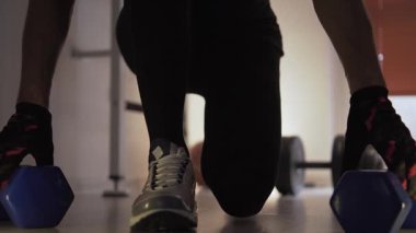 Sporcu diz çöker ve dambılları yere yerleştirir, sonra onları tekrar kaldırır. Arka planda onun yanında yerde büyük bir dambıl var. Spor salonunda ve antrenman yapıyor.