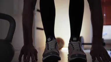Spor ayakkabılı bir sporcu, vücudunu esnetiyor ve gevşetiyor. Önünde bir kettlebell, arkasında da yerde bir dambıl var. Spor salonundaydı ve...