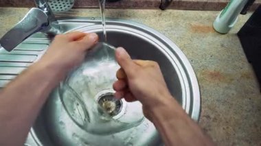 Mutfak lavabosunda güçlü erkek elleri, gümüş renginde, cam kaseyi süngerle yıkıyor. Kase cömert bir su akıntısıyla yıkanır. Birinci kişiden üst görünüm