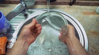 Bir adam gümüş bir mutfak lavabosunda kalın köpükle kaplı bir cam kaseyi yıkıyor. Kase akan suyun altında yıkanır. Birinci şahıstan en iyi görüntü. Büyük yakın çekim.