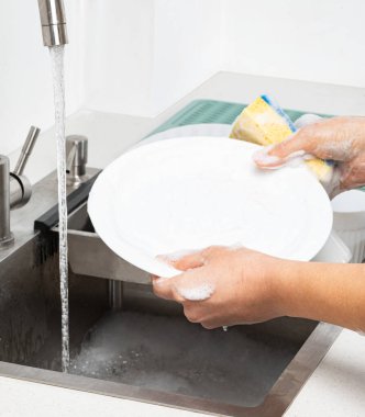 Eller, mutfak lavabosunda akan suyun altında sarı ve mavi bir süngerle beyaz bir tabağı yıkıyor.