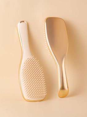 Two hairbrushes, one gold and one cream, are displayed on a matching gold background, creating a stylish and luxurious beauty image
