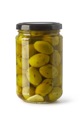Jar containing green olives, garlic cloves, and spices submerged in olive oil, standing on a white background