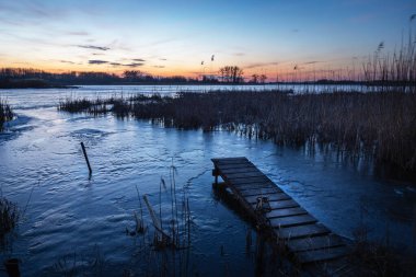 Güneş battıktan sonra ahşap köprüsü olan donmuş mavi bir gölün büyüleyici akşam manzarası..