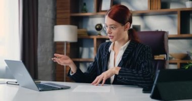 Genç mutlu kızıl saçlı kadın modern ofisteki bilgisayar kullanarak Webinar video eğitiminde konuşuyor. İş kadınının bilgisayarında müşterilerle görüntülü görüşmesi var ve bir şeyler yazıyor. Yavaş çekim.