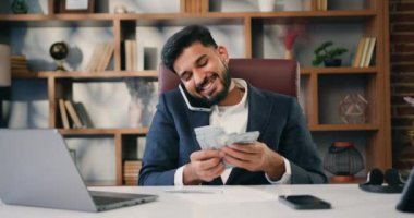 Takım elbiseli iş adamı telefonda konuşuyor ve iş yerinde modern ofiste otururken dolarları sayıyor. Hintli erkek iş adamı. Yavaş çekim.