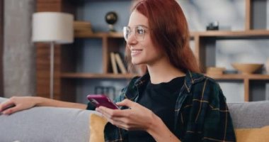 Kızıl saçlı genç bir kadının portresi evde sosyal medya kullanıyordu. Kanepede online alışveriş yapmak için akıllı telefon kullanan güzel bir kız. Yavaş çekim.