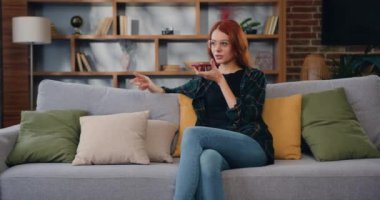Akıllı telefon kaydını kullanan çekici genç bir kadın, oturma odasındaki koltukta otururken sosyal ağda sesli mesajla cevap veriyor. İletişim, internet. Yavaş çekim.