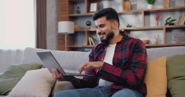 西班牙裔印度人微笑男子远程使用笔记本电脑在网上工作 而坐在沙发上 舒适而时尚的客厅里 虚拟家庭办公室 远程工作 自由职业者在工作 慢动作 — 图库视频影像