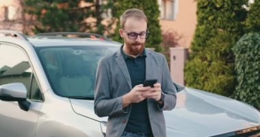 Giysileri içinde yakışıklı beyaz bir adam elinde akıllı bir telefonla sohbet ediyor. Gülümseyen adam modern arabanın yanında sohbet ediyor. Açık hava, internet, dijital, arama iş konsepti. Yavaş çekim.
