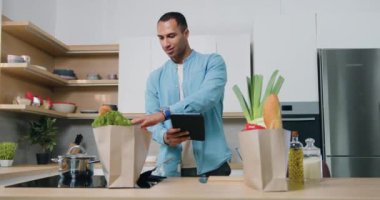 Yakışıklı, bronz tenli, günlük kıyafetlerle gezen adam tablet cihazındaki listeye göre siparişleri kontrol ediyor, mutfaktaki paketleri inceliyor.