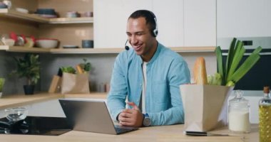 Kulaklıklı Afrikalı bir adam dizüstü bilgisayar kullanıyor ve gülümseyerek görüntülü sohbet yoluyla iletişim kuruyor. Webinar, video konferans, iletişim, e-ticaret, konsept. Yavaş çekim.
