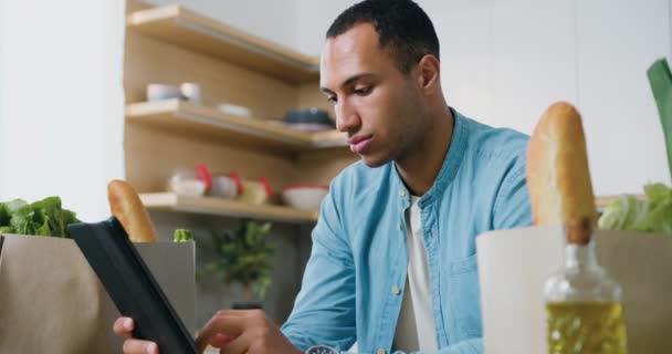 アフリカ系アメリカ人の男性がモダンなキッチンに座り タブレットコンピュータで良いニュースを読んでいます 感情的な幸せな男は 自宅のオフィスでタブレット上の勝利を祝う スローモーション — ストック動画