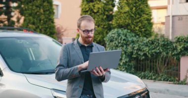 Beyaz, orta yaşlı, güzel bir adam resmi kıyafetlerle siyah arabanın önünde dizüstü bilgisayar kullanarak kentte duruyor. Yavaş çekim.