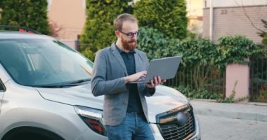 Ceketli, beyaz, yakışıklı bir iş adamı dışarıda, arabanın yanında dizüstü bilgisayar kullanıyor. Duygusal açıdan mutlu bir adam dışarıda laptopta zaferi kutluyor. Yavaş çekim.