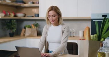 Kendine güveni tam bir kadın portresi, günlük tarz kıyafetleri içinde, kadın serbest, evde dizüstü bilgisayarın yanında duruyor, kameraya bakıp gülümsüyor ve başparmaklarını gösteriyor. Yavaş çekim.