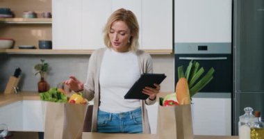 Mutfak masasının başında duran gülümseyen genç kadın tablet dijital tableti tutuyor dengeli beslenme için sağlıklı tarifler arıyor, internette öğle yemeği için ürünler sipariş ediyor. Yavaş çekim.