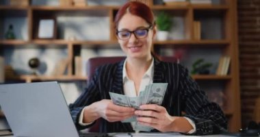 Takım elbiseli neşeli CEO kadın modern ofiste oturmuş nakit para sayıyor. İş kadını, yığınla nakit para, dolar, kar, maaş, iş yeri. Yavaş çekim.