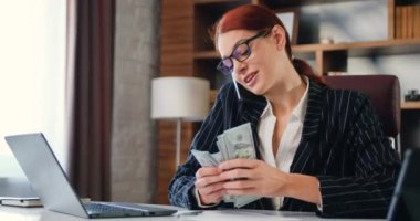 Takım elbiseli bir iş kadını, iş yerinde modern bir ofiste otururken telefonla konuşuyor ve dolarları sayıyor. Ünlü işyeri sahibi..
