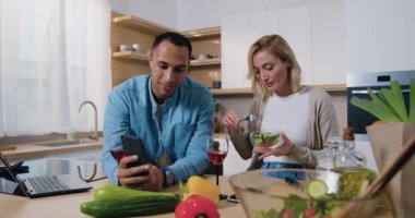 Mutlu aile evde birlikte vakit geçiriyor. Adam telefonda bir kadına bir şey gösteriyor, kadın gülümsüyor ve salata yiyor. Modern mutfakta telefonu kullanan çok kültürlü genç bir aile. Yavaş çekim.