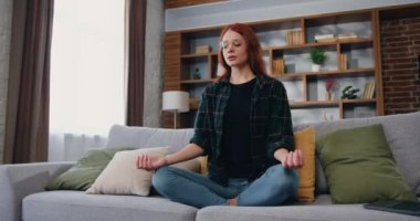 Pratik, iç ahengi ve dengeyi hisseder. Stres yok. Çekici kızıl saçlı beyaz kadın Lotus pozisyonunda yoga yapıyor ve meditasyonun gözlerini kapıyor. Evdeki koltukta oturuyor. Yavaş çekim.