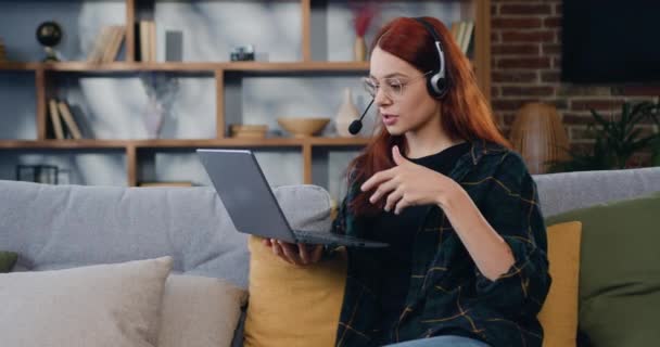 戴耳机的年轻女人 会说话的视频电话 坐在家里的沙发上 在线教育 远程培训 商务会议 网络研讨会 慢动作 — 图库视频影像