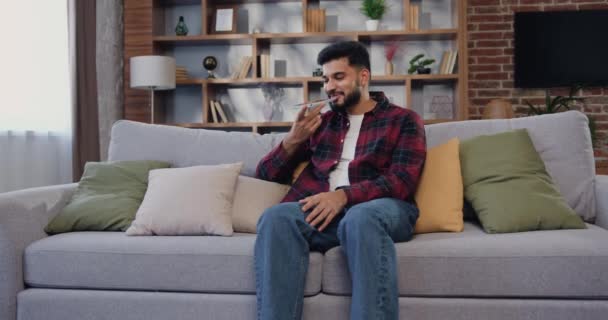 光のリビングルームでソファに座っている間 ソーシャルネットワーク内のオーディオメッセージに応答するスマートフォンの録音を使用して魅力的なインド人男性 インターネット スローモーション — ストック動画
