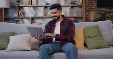 Genç, güzel, yetişkin Hintli bir adam rahat bir oturma odasında elinde dizüstü bilgisayarla oturuyor. Yaratıcı İspanyol erkek sosyal medyayı kontrol ediyor, internette geziniyor. Yavaş çekim.