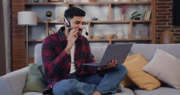 自宅のソファに座ってラップトップを使用してヘッドフォンで笑顔の男 ヒスパニック系男性で現代のヘッドフォンでソファの上にリビングルームに座ってビデオ通話上のラップトップ — ストック動画