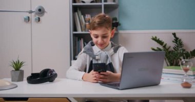 Akıllı telefonu elinde tutan sevimli çocuk mobil uygulamalar kullanarak eğleniyor, evdeki oturma odasındaki masada dizüstü bilgisayarın yanında oyunlar oynuyor. Yavaş çekim.