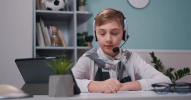Concentrated little 19-10s kid boy wearing wireless headphones listening educational lecture in online school. Boy writes in notebook and looks at computer screen. Slow motion.