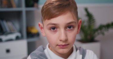 Portrait of the cute little blonde boy sitting in the living room table looking at the camera. Face eyes smiling contemplative child. Close Up. Slow motion.