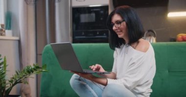 Young woman in glasses sits at on sofa at laptop computer working at home. Female freelancer or student listens to an online conference in the kitchen. Slow motion.