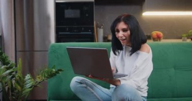 Woman reading good news on laptop screen at home. Winner amazed female sits in kitchen celebrates victory wins online sales discounts internet auction bid success. Slow motion.