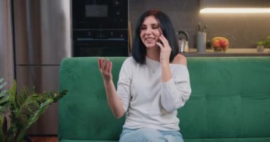 Young smiling woman sitting on sofa and talking smartphone at home. female freelancer using cellphone discussing business project with colleague. Kitchen on background. Slow motion.