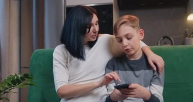 Mother and son sitting on sofa at home. Adult mom teaching cute kid son learning using smartphone funny apps or playing mobile games using apps for tech education entertainment in kitchen. Slow motion