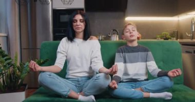 Güzel anne ve çocuk oğul birlikte yoga yapıyorlar. Lotus pozu veriyorlar ve birlikte meditasyon yapıyorlar. Evdeki kanepede oturuyorlar. Sağlıklı Yaşam Tarzı, Yaşam Dengesi. Yavaş çekim.