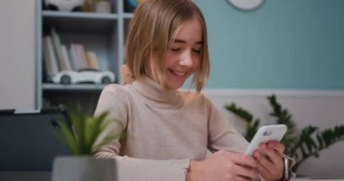 Smiling cute preteen girl use mobile phone sitting by table in living room, getting good news, smiling, laughing, looking at screen. Slow motion