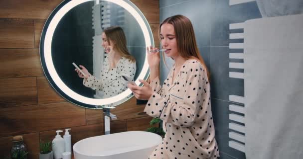 Młoda Uśmiechnięta Kobieta Używa Smartfona Pracy Lub Rozrywki Podczas Mycia — Wideo stockowe