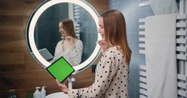 Happy woman brushing teeth and looking on tablet green screen pc in modern bathroom. Beautiful girl in a white terry dressing gown with telephone addiction. Slow motion.