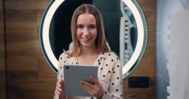 Caucasian smilling woman standing in bathroom and holds a digital tablet pc. Attractive girl looking at camera in bathroom. Beauty and style concept. Slow motion.