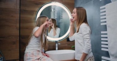 Caucasian young and child sisters brushing teeth looking at each other smiling. Smilling woman and kid taking care of dental health in the morning in bathroom. Slow motion.