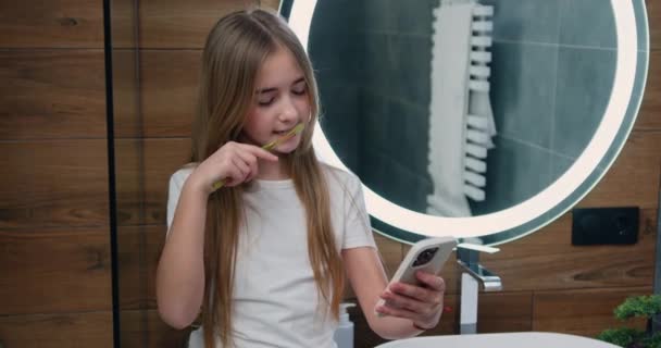 Cute Little Girl Brushing Teeth Using Smartphone Checking Social Networks — Stock Video