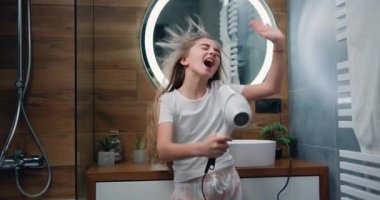 Beautiful blond little girl in bathrobe singing a song and dancing with a hair dryer. Child sings and dances in a shower after taking shower in bathroom. Slow motion.