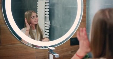 Little attractive girl brushing her hair and assembles in a pigtail in the bathroom. The girl looks directly into the mirror. Slow motion.