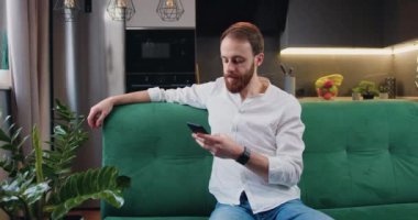 Portrait of man sitting on the sofa using phone in the modern kitchen texting message scrolling tapping technology. Caucasian male relaxing sitting on couch at home Slow motion.