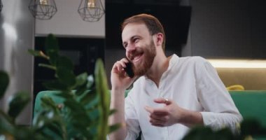 Young man is sitting at home on the couch and smiling talking on a cell phone. Male freelancer using cellphone discussing business project with colleague. Kitchen on background. Slow motion.