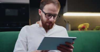 Young man using digital tablet sitting on sofa at home. Male holding laptop looking at screen reading e-book app online relaxing on leisure with wireless device work online. Slow motion.